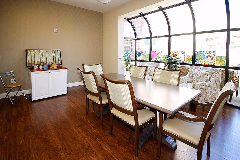A bright dining area with a rectangular table surrounded by six wooden chairs with cream-colored cushions. The room has wooden flooring and beige patterned wallpaper. A white cabinet with a framed picture and small decorative birdhouses sits against one wall. Large arched windows with colorful flower artwork on the lower panes let in natural light, and a cozy seating area with floral-patterned armchairs is visible near the windows.