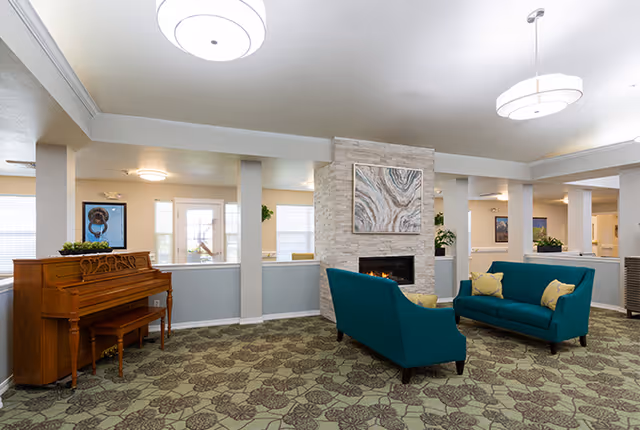 A spacious senior living facility common area with two teal sofas facing a modern fireplace set in a stone wall. Above the fireplace is an abstract painting. To the left, there is a wooden piano with a small bench. The room has patterned carpet flooring, white walls with gray wainscoting, and ceiling lights providing bright illumination.