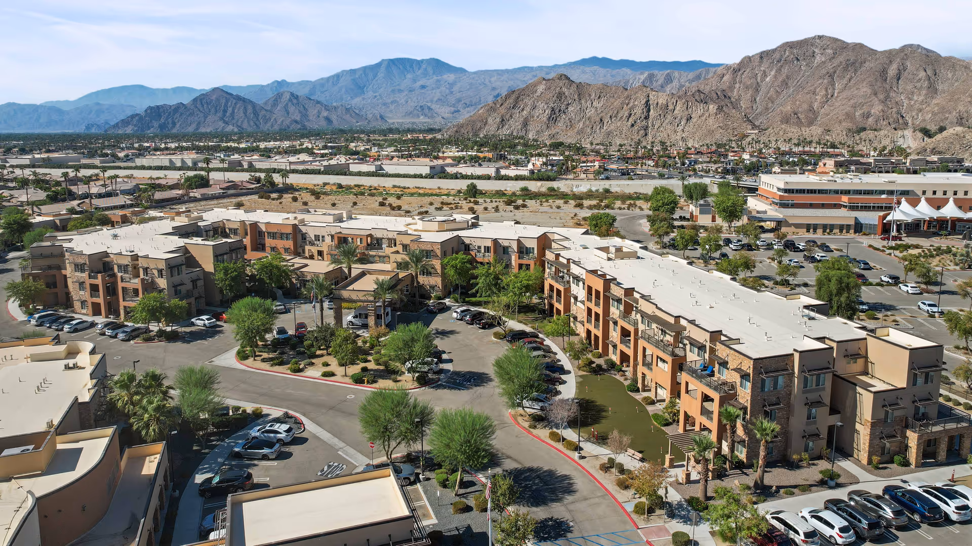 Aerial view of The Palms At La Quinta Gracious Retirement Living facility showing multiple connected buildings with parking lots, landscaped areas with trees and shrubs, and surrounding mountainous terrain under a clear sky.