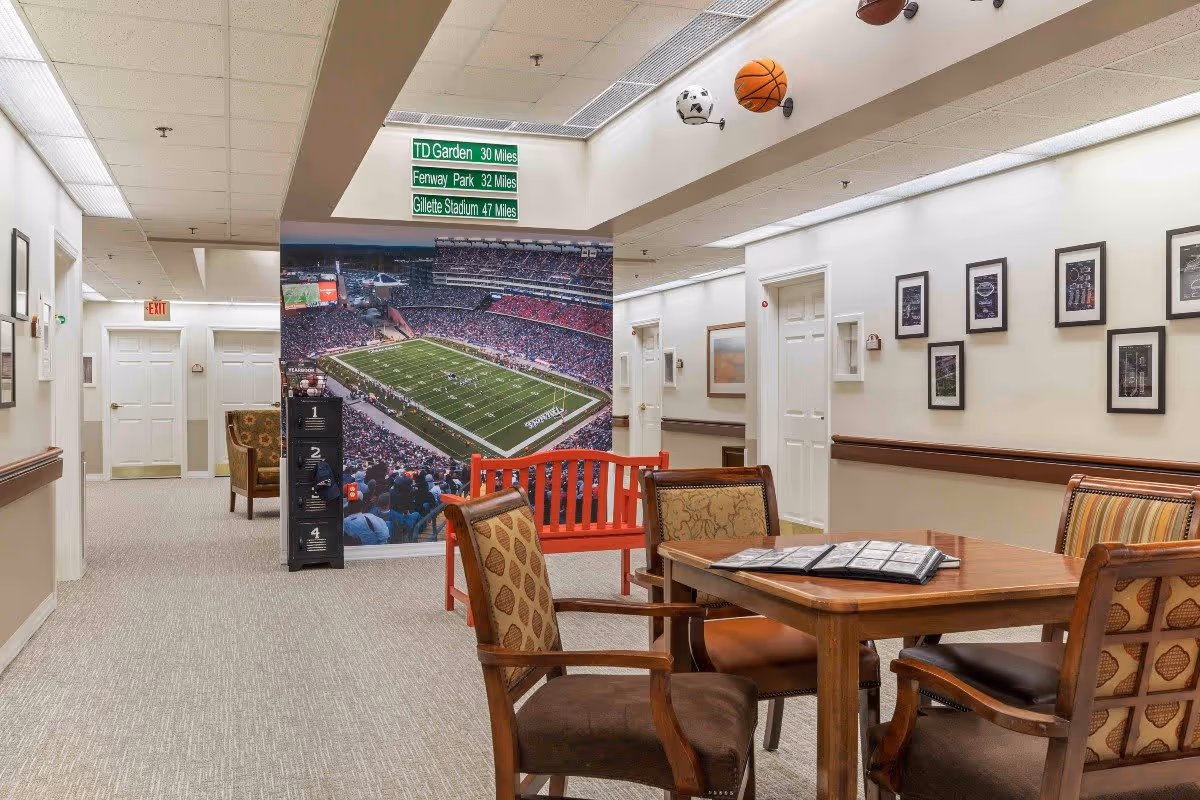 Interior hallway of The Atrium at Drum Hill featuring a seating area with a wooden table and four chairs. On the wall is a large mural of a football stadium filled with spectators. Above the mural, a sign indicates distances to TD Garden, Fenway Park, and Gillette Stadium. The hallway has beige walls, carpeted floors, framed pictures, and sports-themed decor including mounted balls on the ceiling.