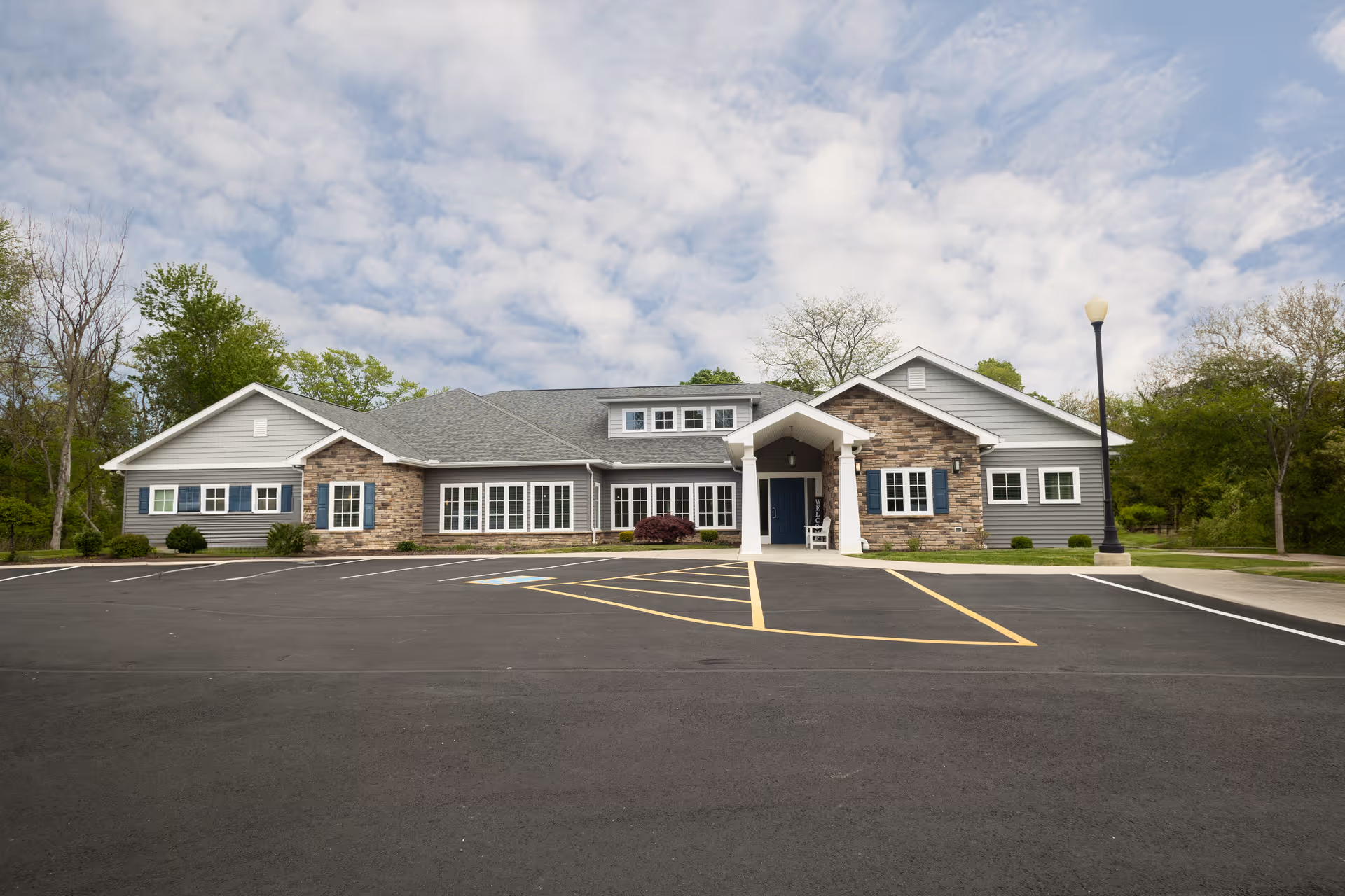 Front exterior view of a single-story senior living community building with a combination of stone and gray siding, multiple windows, a covered entrance with white pillars, a parking lot with marked spaces, and a lamppost on the right side. Trees and greenery surround the building under a partly cloudy sky.