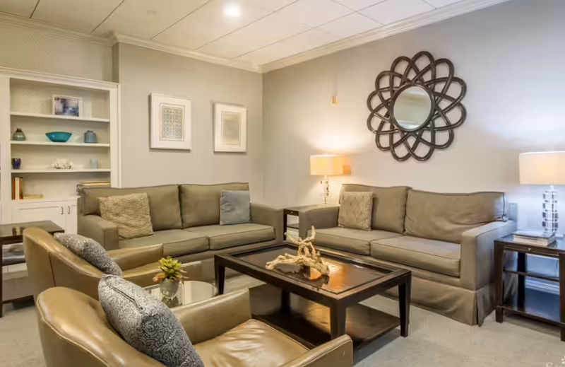 A cozy living room with two gray sofas, two brown leather armchairs, a dark wooden coffee table with decorative driftwood, two side tables with lamps, a round mirror with a decorative frame on the wall, and a built-in white bookshelf with decorative items.