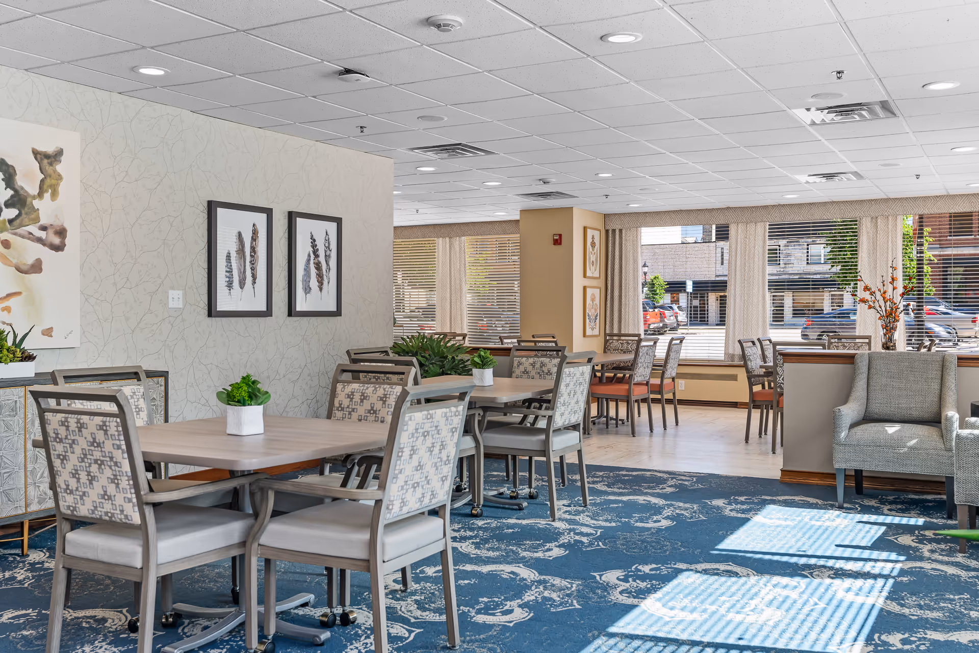 A bright and spacious dining area with multiple tables and chairs arranged neatly. The room features large windows with blinds allowing natural light to fill the space. There are framed artworks on the walls and small potted plants on the tables. A comfortable armchair is positioned near a half wall, and the floor is covered with a blue patterned carpet and light wood flooring near the windows.