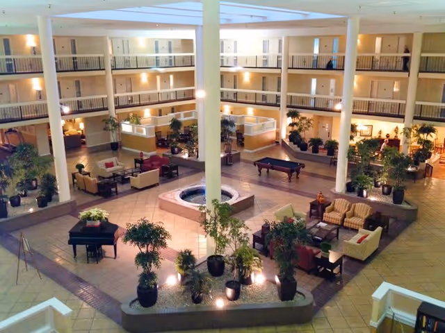 Spacious indoor atrium area of a senior living facility with multiple seating arrangements including sofas and chairs, a central water fountain, numerous potted plants, a piano, and a pool table. The atrium is surrounded by multiple floors with railings and doors leading to rooms.
