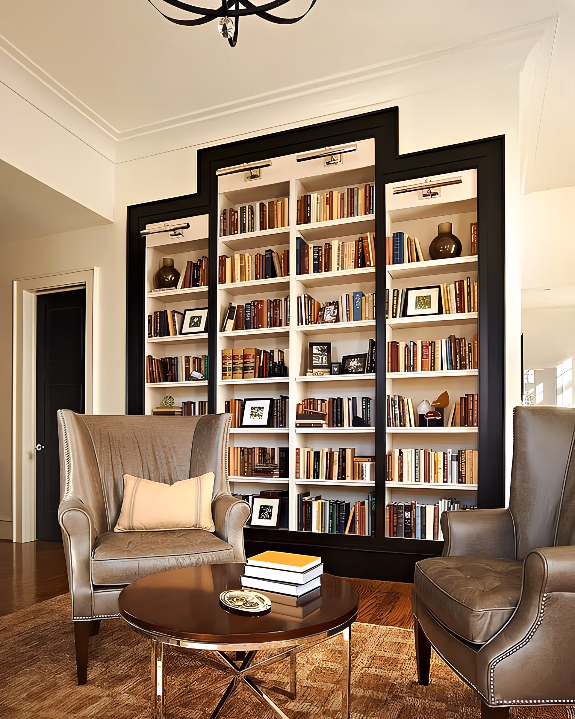 A cozy living room area featuring two brown leather armchairs with a beige pillow on one, a round wooden coffee table with a stack of books and a decorative item, and a large built-in bookshelf filled with books and framed photos, framed by a black border.