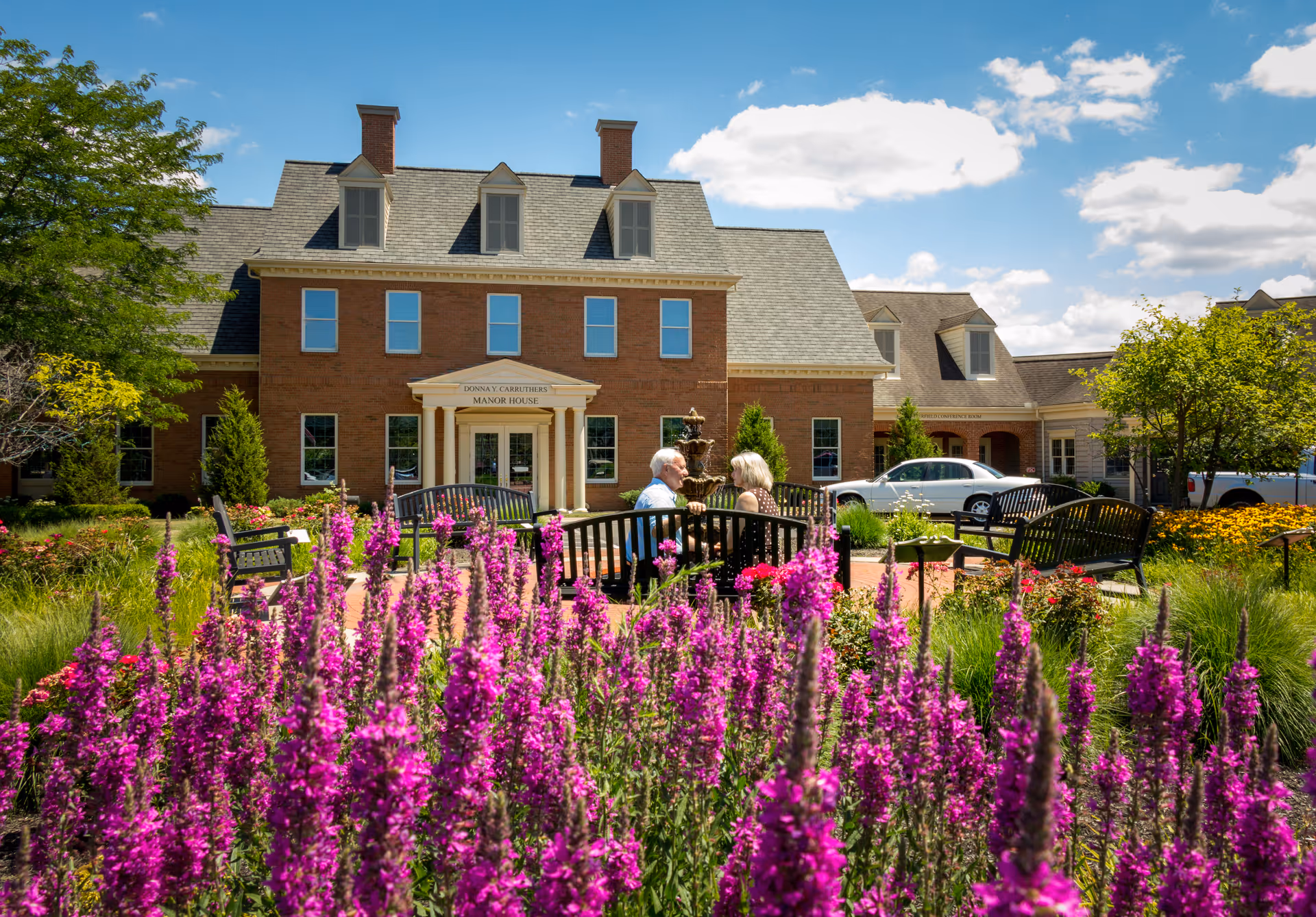 Brick manor-style senior living building with a landscaped front garden of vibrant purple flowers, benches, and people seated near the entrance.