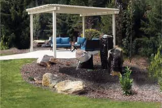 Outdoor garden area with a white pergola covering blue cushioned seating. There are tree stumps and rocks arranged in a mulched bed surrounded by green grass and tall evergreen trees in the background.