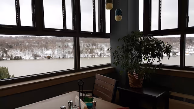 View from inside a room with large windows overlooking a snowy landscape with a river and trees. Inside, there is a table with condiments and a chair, and a potted plant on a small table near the window.