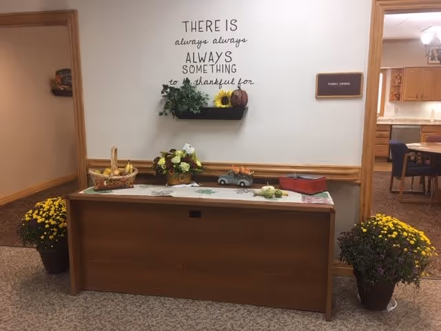 A hallway corner in a senior living facility with a wooden cabinet decorated with a basket of bananas, a flower arrangement, a small toy truck with pumpkins, and a red box. Above the cabinet is a wall decal that reads 'There is always always always something to be thankful for' with a small shelf holding a plant, a sunflower, and a pumpkin. Two potted yellow and purple flowers are placed on the floor on either side of the cabinet. A sign on the wall to the right reads 'Family Dining' and an open doorway reveals a dining area with tables and chairs.