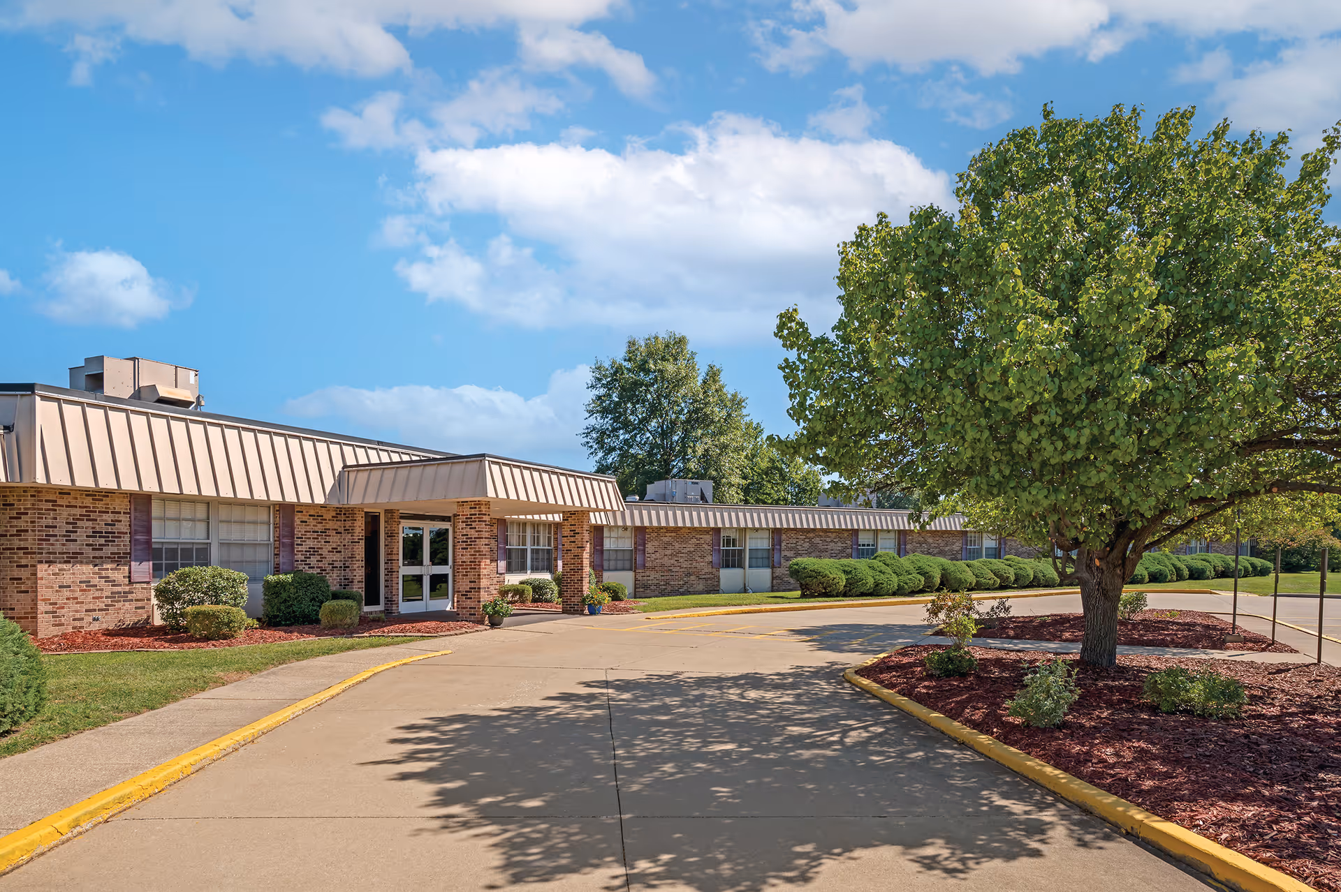 Exterior view of a single-story brick building with a metal roof, surrounded by neatly trimmed bushes and a large tree. The sky is blue with some clouds, and there is a paved driveway leading to the entrance of the building.