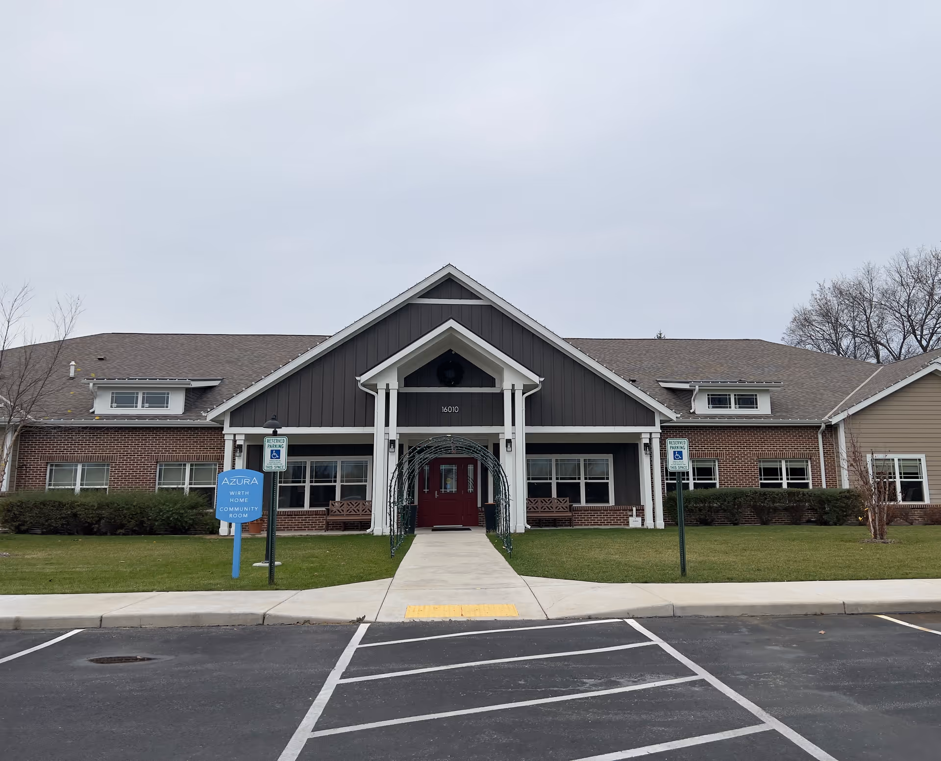 Front exterior of a single-story brick and siding senior living building with a central covered entrance, walkway, lawn and parking spaces.