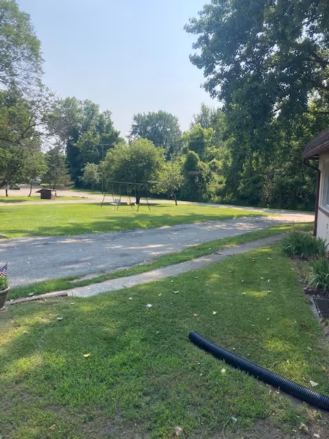 Outdoor area with a grassy lawn, a gravel driveway, and a swing set in the distance surrounded by trees under a clear sky.
