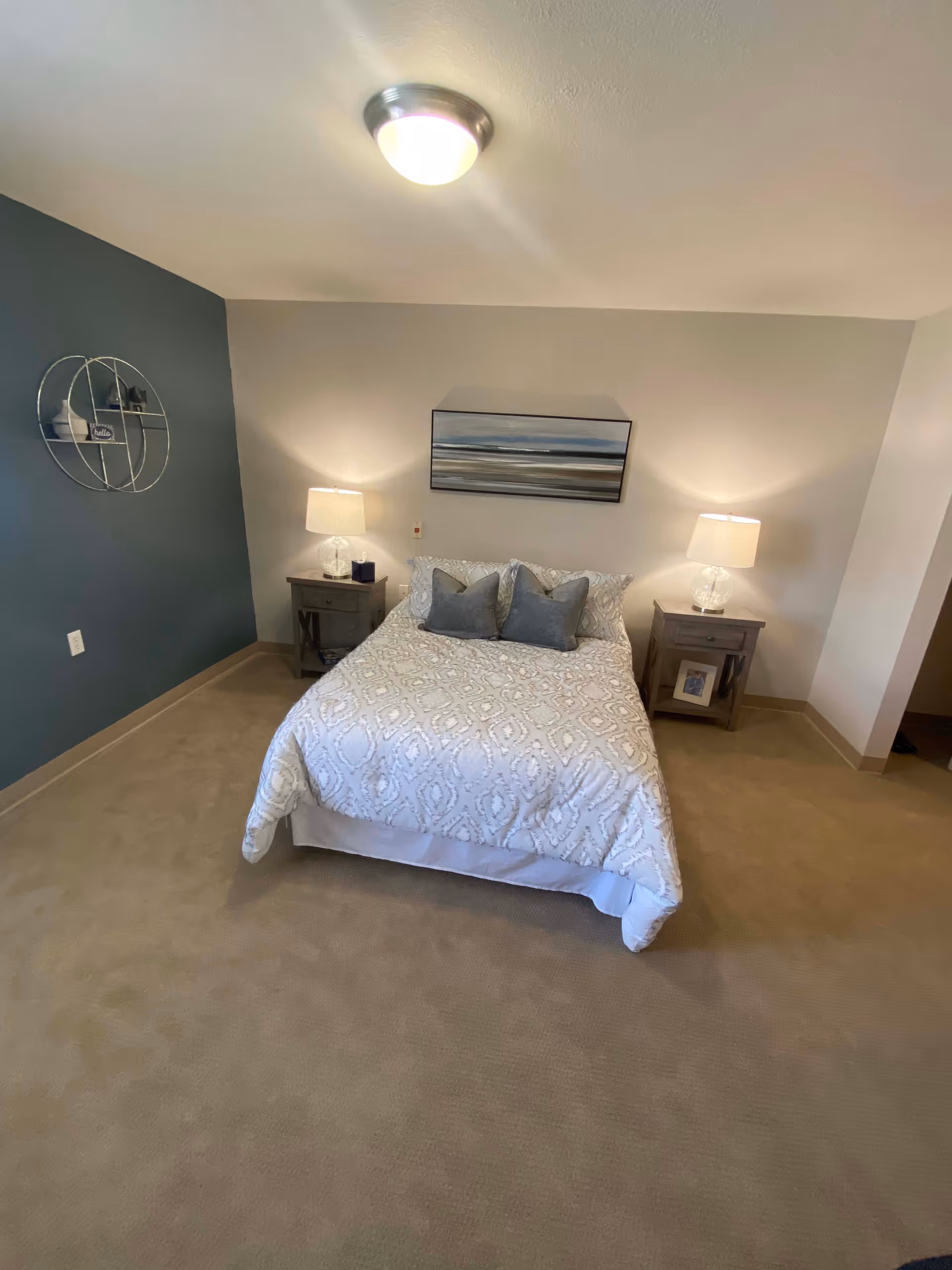 A neatly made bed with a white patterned comforter and two gray pillows in a bedroom. There are two wooden nightstands on either side of the bed, each with a lamp. A framed picture hangs above the bed, and a circular wall shelf with decorative items is mounted on the dark accent wall to the left. The room has beige carpet and neutral-colored walls.