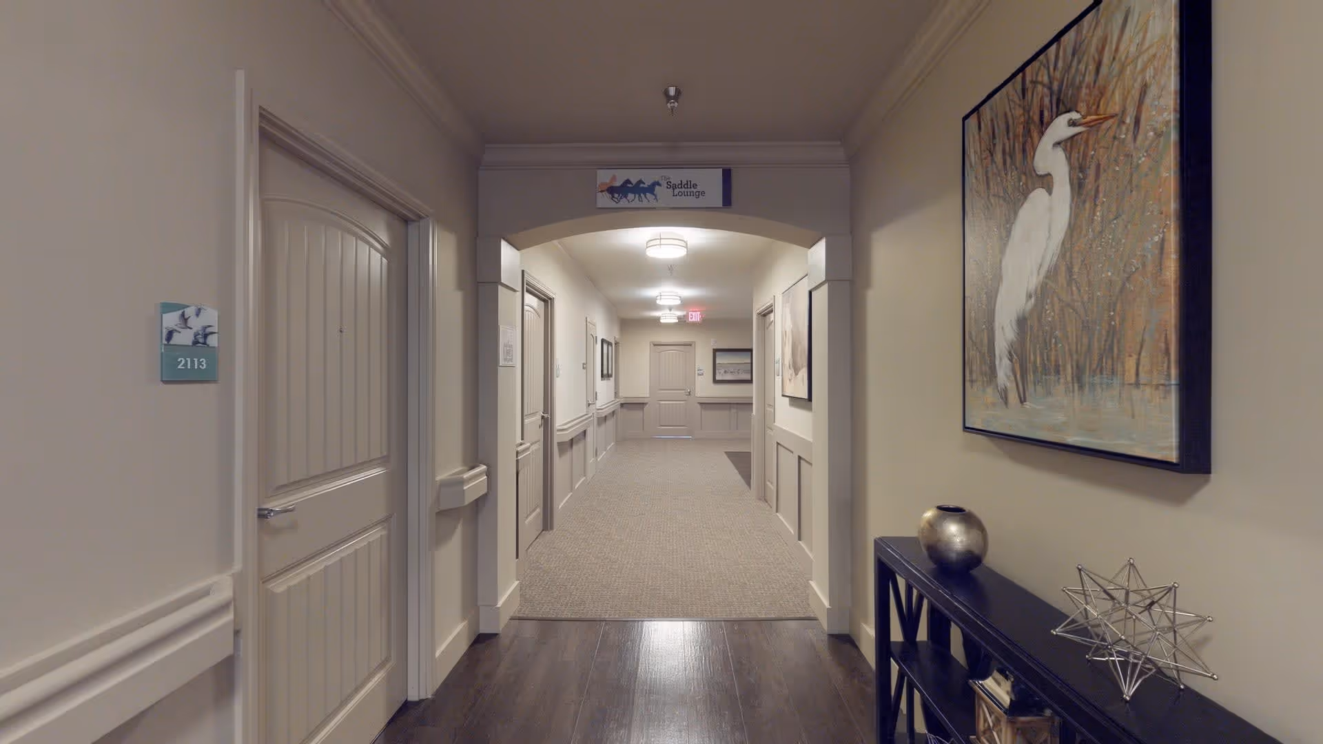 A well-lit hallway in a senior living facility with beige walls and carpeted flooring. There are several closed doors along the hallway, a sign above the archway reading 'Saddle Lounge,' and a console table on the right side with decorative items including a metallic vase and a geometric sculpture. A large painting of a white bird in a natural setting hangs above the console table.