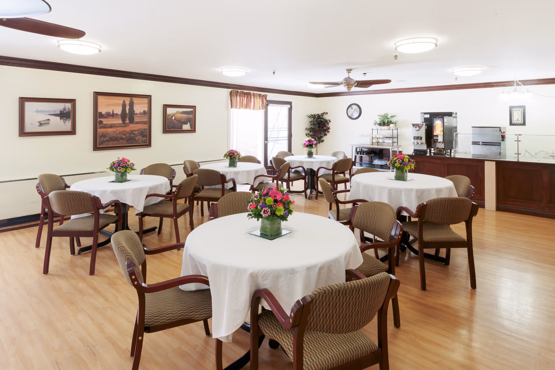 A bright dining room with several round tables covered with white tablecloths, each adorned with a colorful floral centerpiece. The room has wooden floors, beige walls with framed landscape paintings, and multiple chairs around each table. In the background, there is a counter area with coffee and beverage machines, a clock on the wall, and a potted plant near a window with a curtain.
