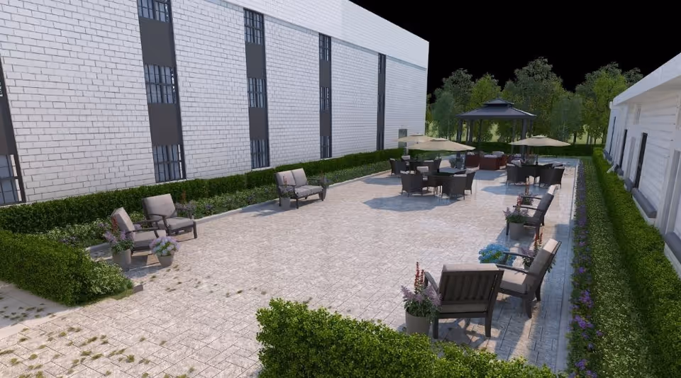 Outdoor patio area with multiple seating arrangements including chairs, tables with umbrellas, and a gazebo surrounded by greenery and hedges next to a building.