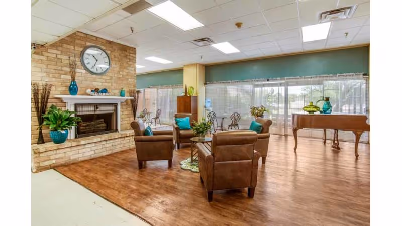 A spacious living area with wooden flooring featuring a brick fireplace with a clock above it, four brown armchairs arranged around a small table with a plant centerpiece, large windows with blinds letting in natural light, a wooden piano with decorative vases on top, and additional seating with metal chairs near the windows.