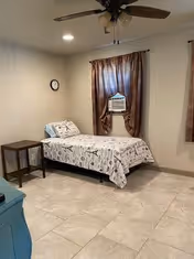 Simple bedroom with a single bed, nightstand, window air conditioner beneath curtains, ceiling fan, wall clock and tiled floor.