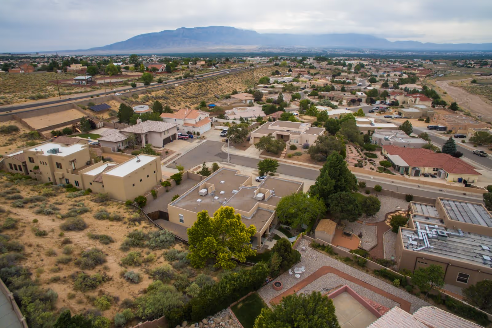 Aerial view of a suburban neighborhood with houses, roads, and scattered trees, set against a backdrop of mountains under a cloudy sky.