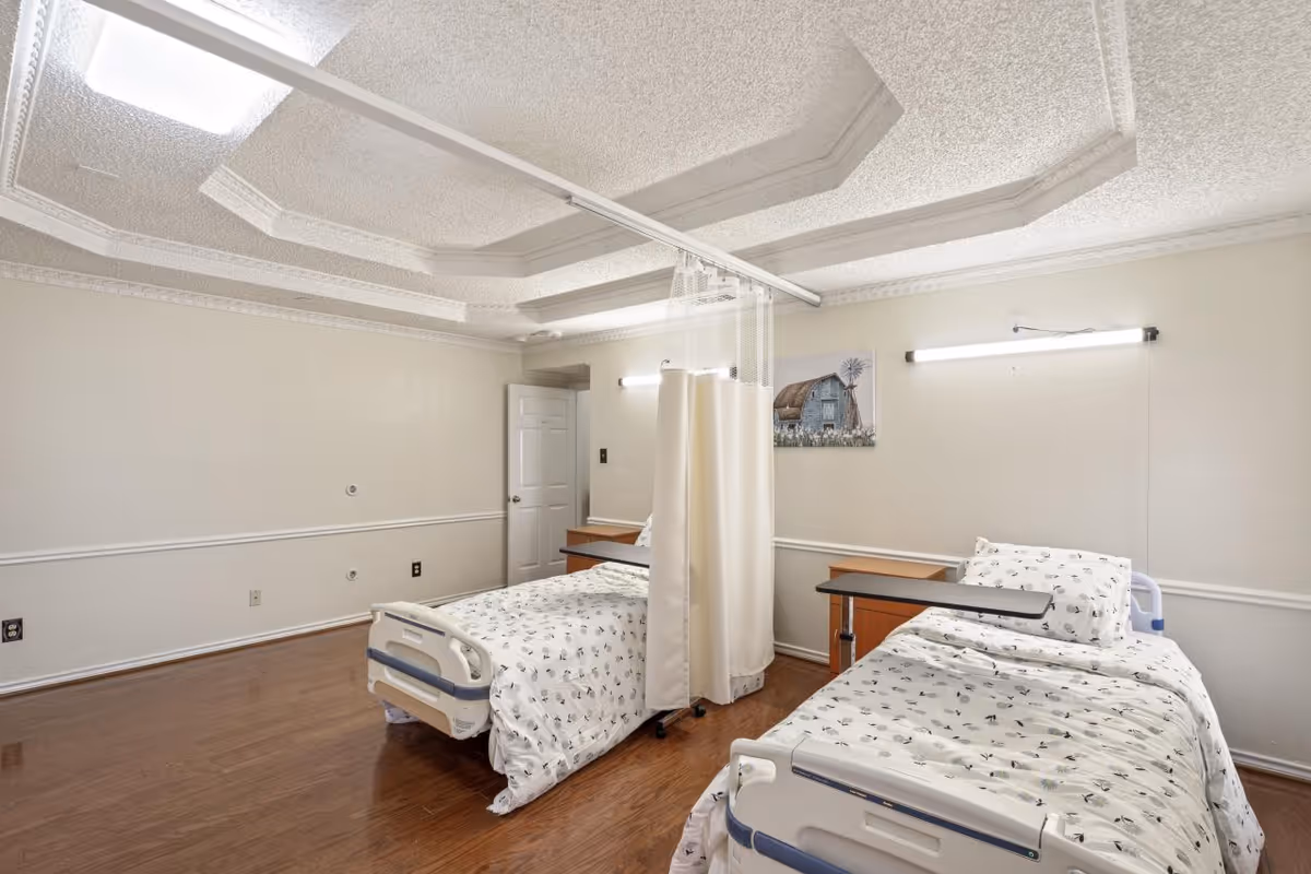 A clean, well-lit room with two hospital-style beds separated by a privacy curtain. Each bed has white bedding with a subtle pattern, and there are small wooden bedside tables next to each bed. The room features a decorative ceiling, hardwood floors, and a painting of a barn on the wall.