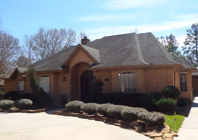 Exterior view of a single-story brick building with a dark shingled roof, arched entrance, and several windows with white blinds. The building is surrounded by neatly trimmed bushes and landscaping with a concrete driveway in front. Trees without leaves are visible in the background under a clear blue sky.