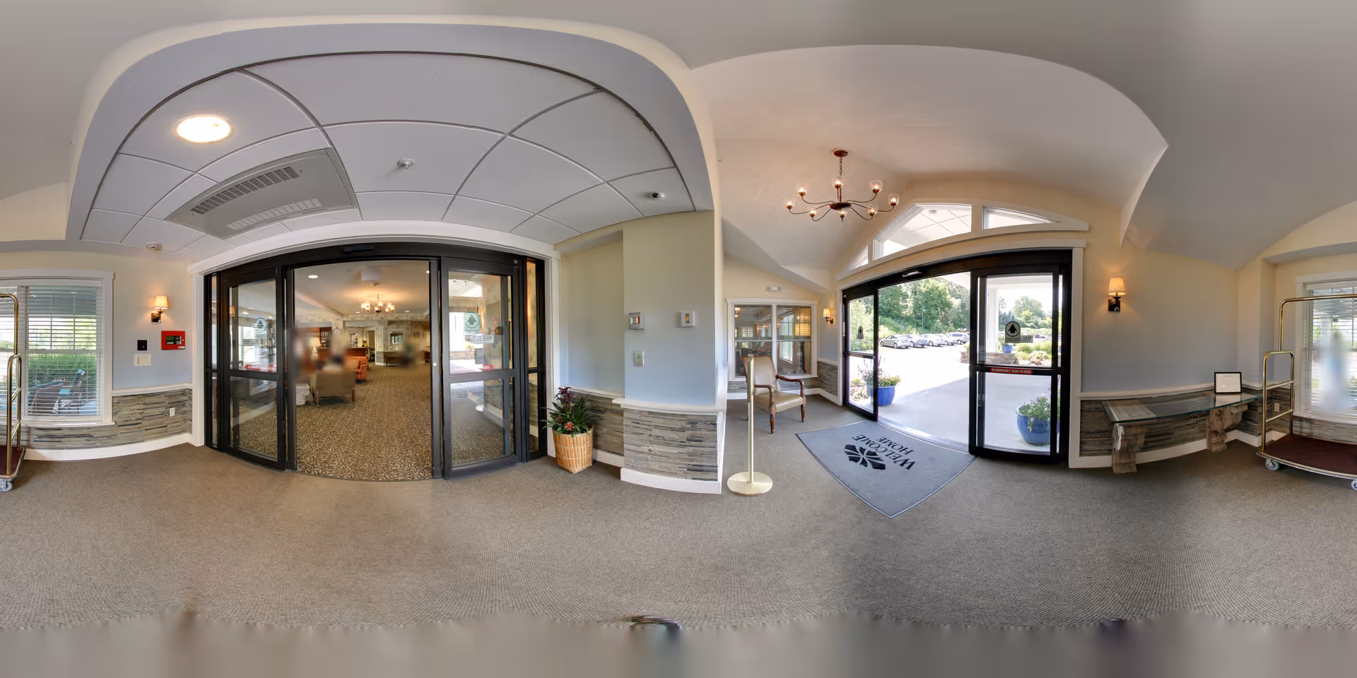 Entrance lobby of a senior living facility with glass sliding doors, seating, chandeliers, and a welcome mat.