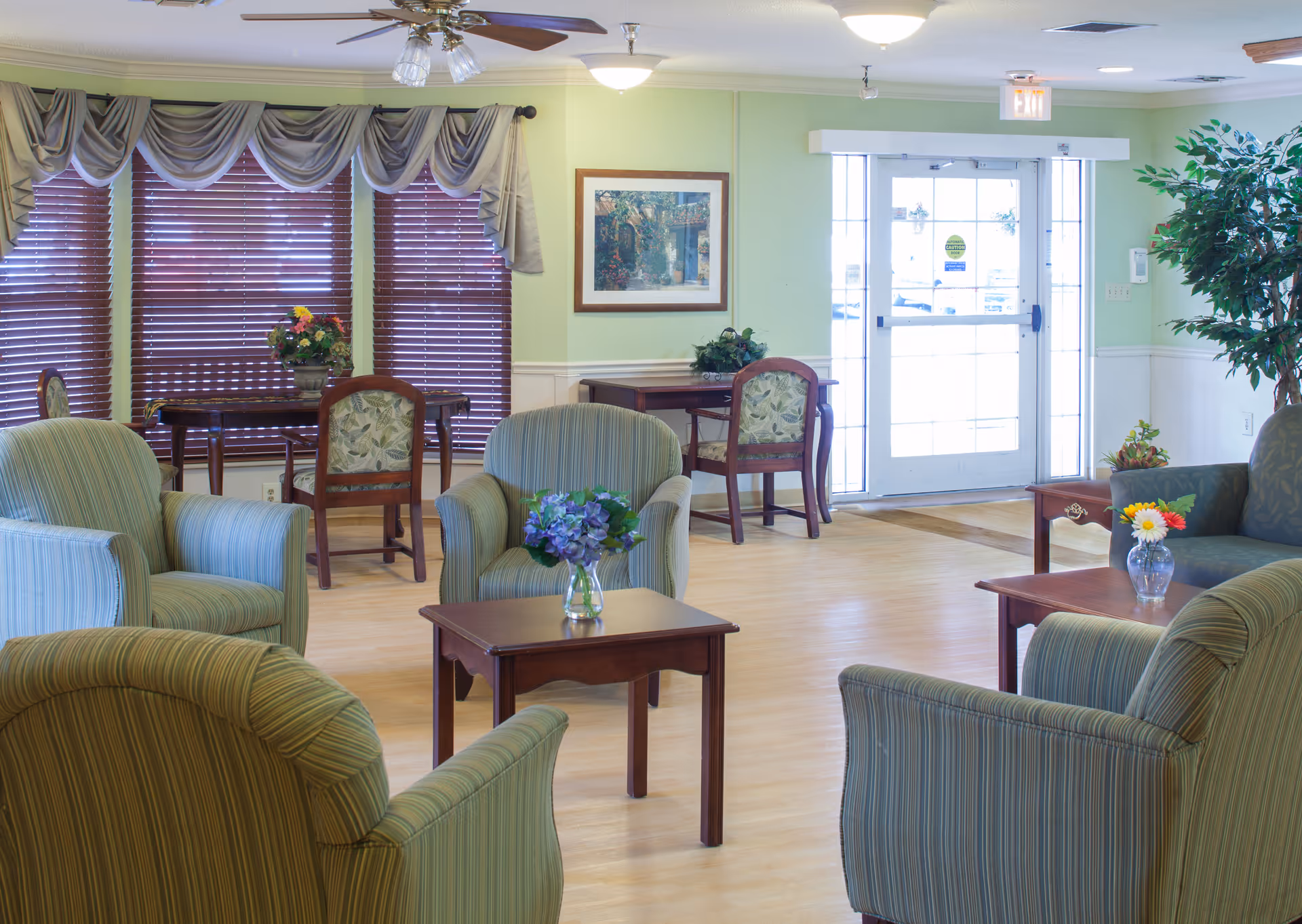A bright and welcoming common area with several striped upholstered armchairs arranged around wooden coffee tables, each table adorned with a small vase of flowers. In the background, there are wooden tables and chairs near windows with closed blinds and decorative valances. A framed picture hangs on the light green wall, and a glass door with sidelights leads outside. A large potted plant is visible on the right side of the room.