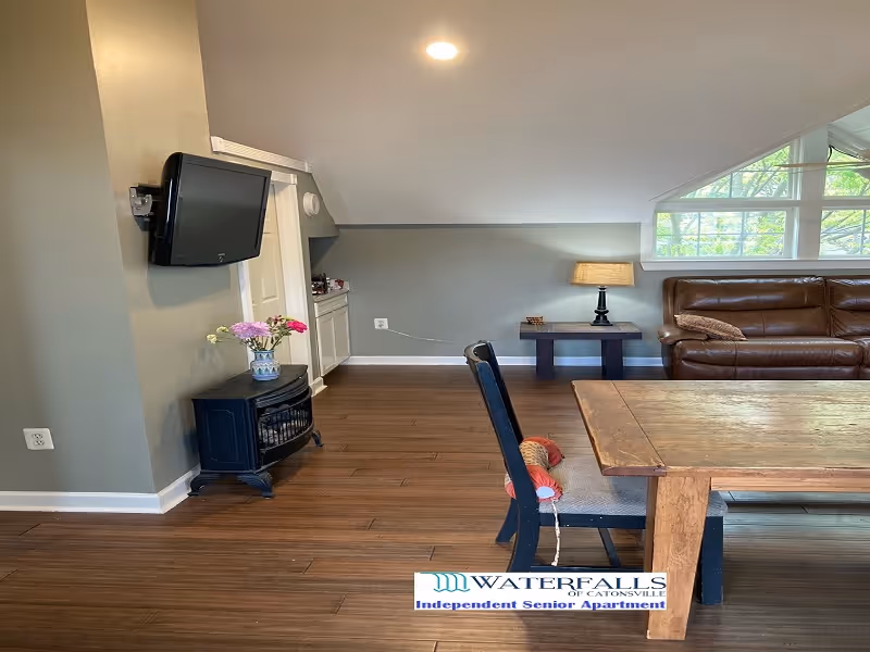 Open living area with a wooden dining table and chairs, a leather sofa by a window, a wall-mounted TV, and a side table with a lamp.