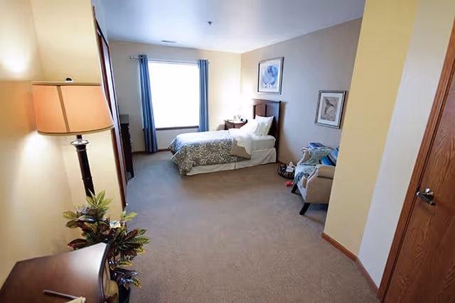 A spacious bedroom with a single bed against the far wall, a window with blue curtains, a bedside table with a lamp, a comfortable armchair with a blanket, framed artwork on the walls, a floor lamp, and a plant near a wooden dresser.