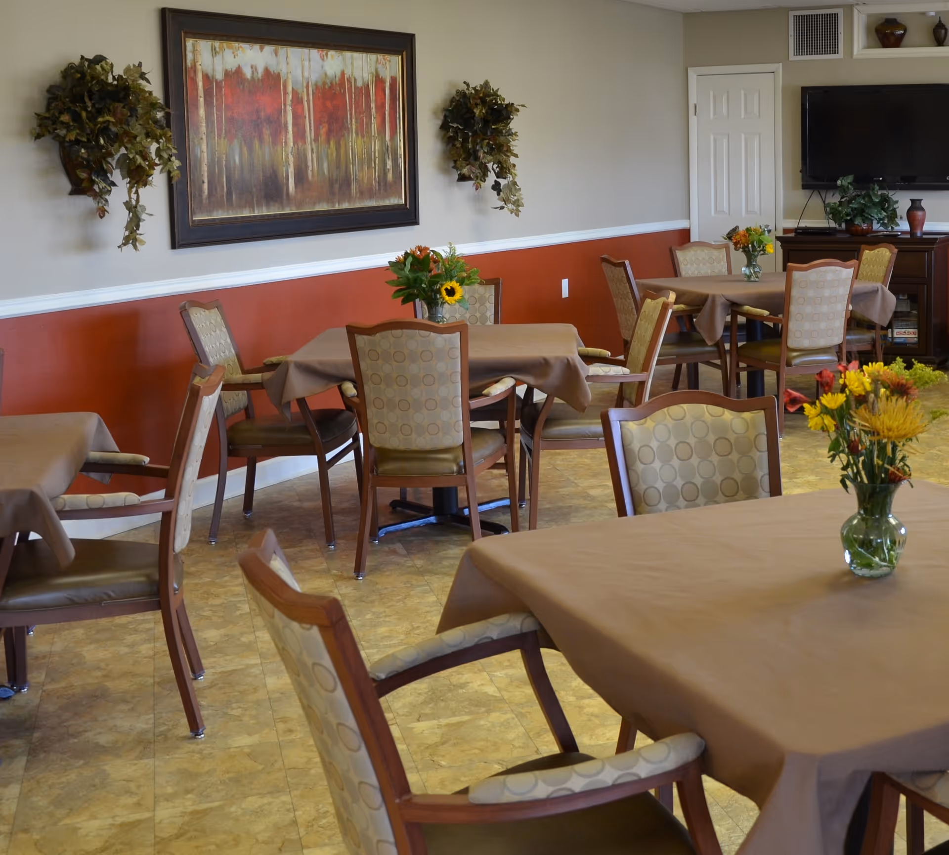 A dining room with several tables covered with brown tablecloths and surrounded by cushioned chairs with patterned upholstery. Each table has a small vase with colorful flowers. The walls are painted beige and red with a white chair rail, decorated with a large framed painting and two wall-mounted plants. A flat-screen TV is mounted on a cabinet in the background.