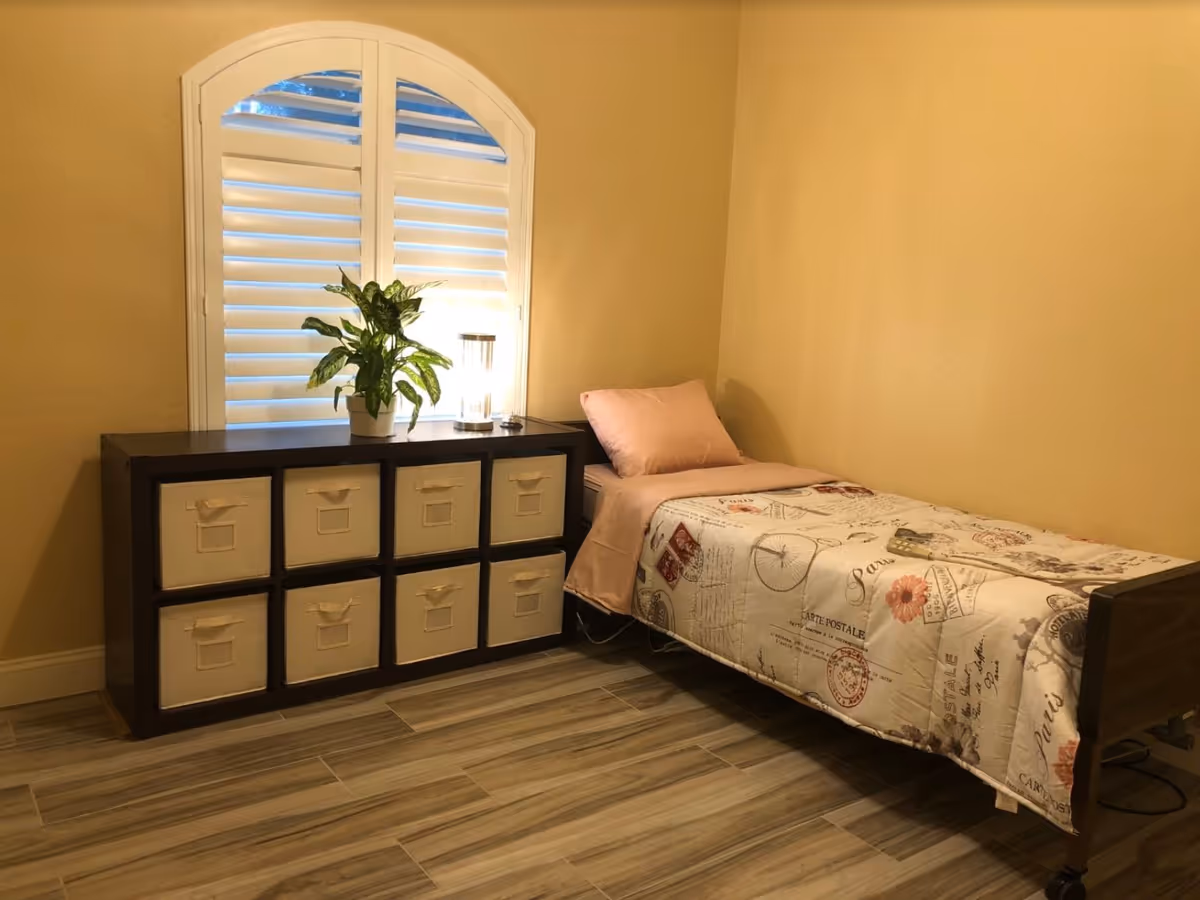 A small bedroom with a single bed covered by a patterned quilt and pink pillow, a cube storage unit topped with a potted plant and lamp, and a shuttered arched window.
