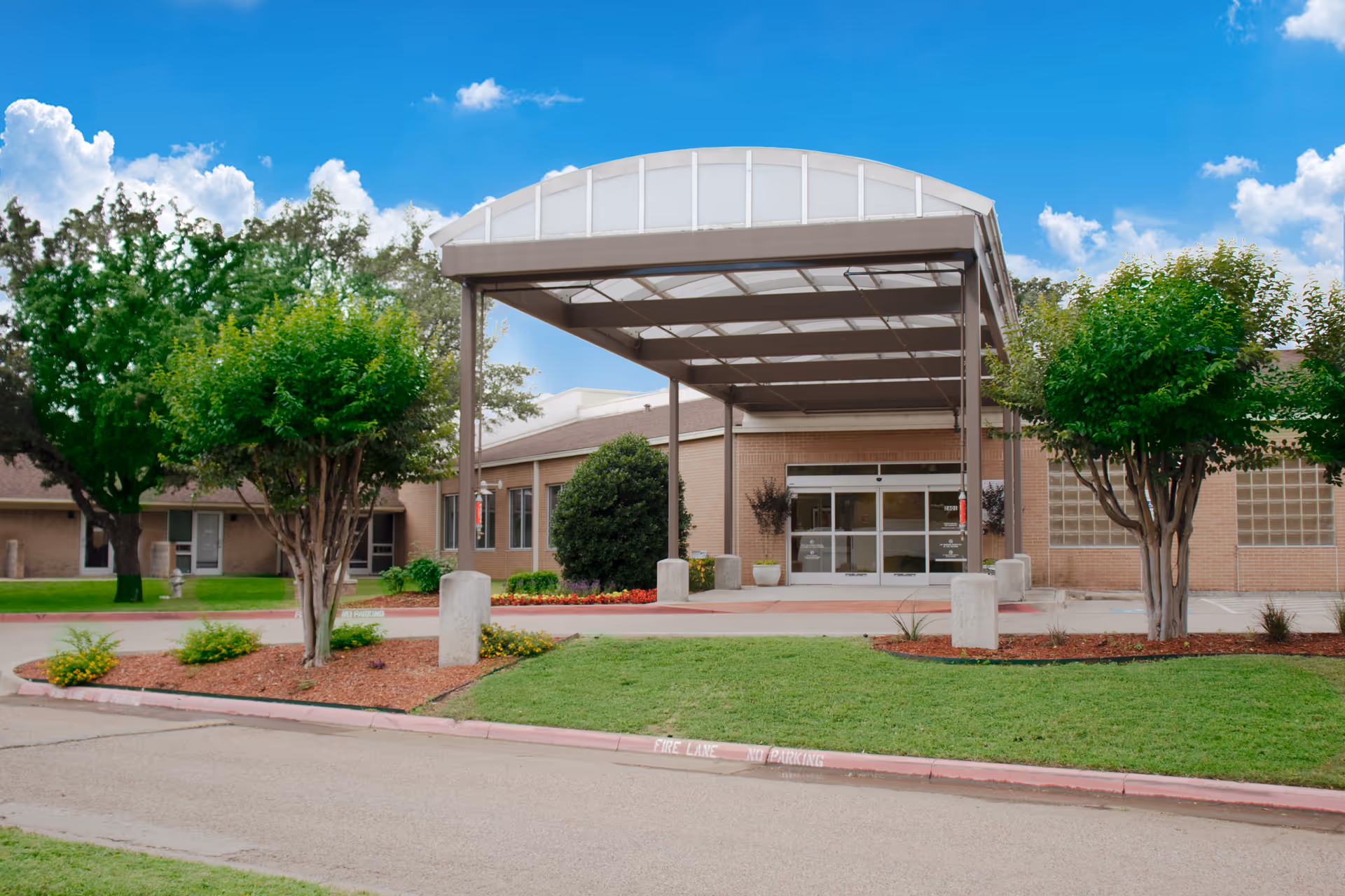 Entrance of Arlington Villas Rehabilitation And Healthcare Center showing a covered drop-off area with a metal canopy, surrounded by green trees and landscaped grass areas under a blue sky with scattered clouds.