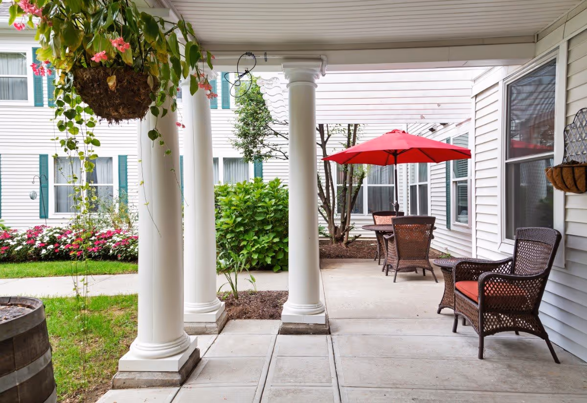 Outdoor patio area at Brighton Gardens of Stamford featuring white columns, wicker chairs with red cushions, a table with a red umbrella, hanging flower baskets, and surrounding greenery with flowering plants and bushes.