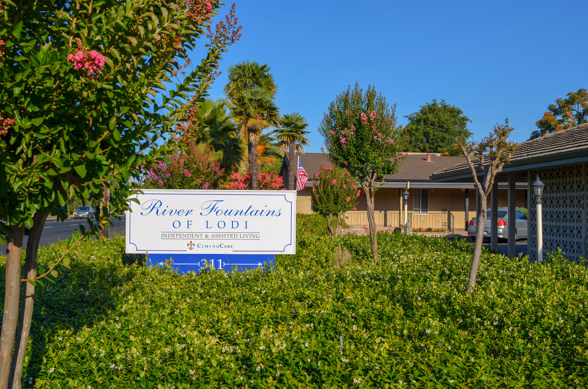 Outdoor view of River Fountains of Lodi senior living facility sign surrounded by green bushes and trees with pink flowers, with a building and parked cars in the background under a clear blue sky.