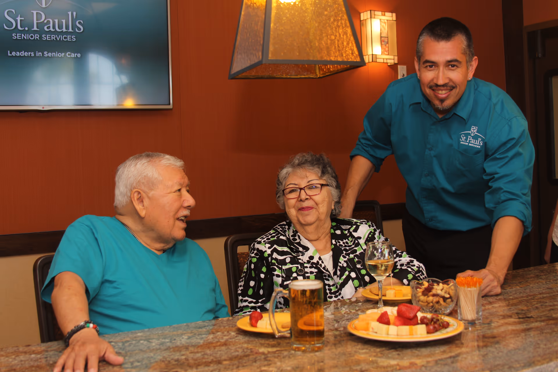 Two elderly residents sit at a dining counter with plates of fruit and drinks while a staff member in a St. Paul's shirt stands behind them.