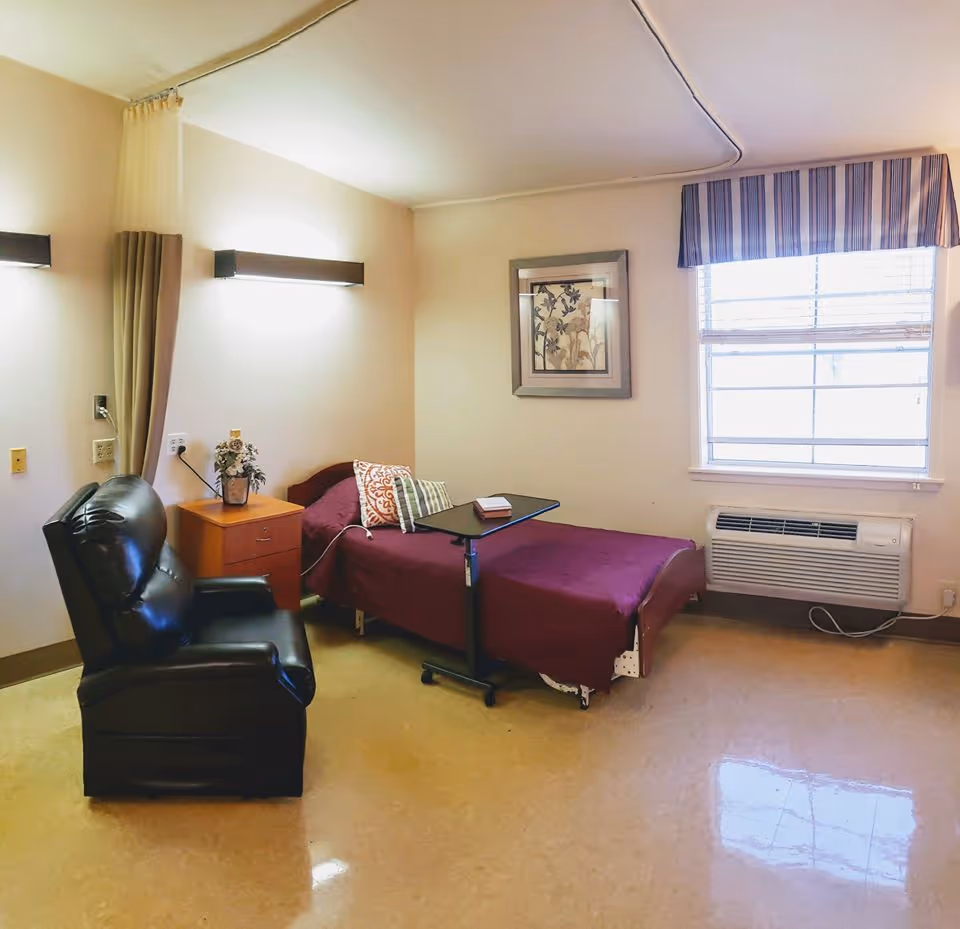 A simple, clean bedroom in a senior care facility featuring a single bed with a maroon bedspread and decorative pillows, a small wooden nightstand with a vase of flowers, a black leather recliner chair, a window with striped valance, and a wall-mounted air conditioning unit below the window.