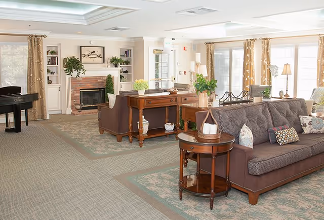 A bright and spacious living room area with large windows covered by patterned curtains, a brown sofa with decorative pillows, wooden side tables with plants and candles, a fireplace with a brick surround and white mantel, built-in shelves with books and plants, and a black grand piano in the corner.