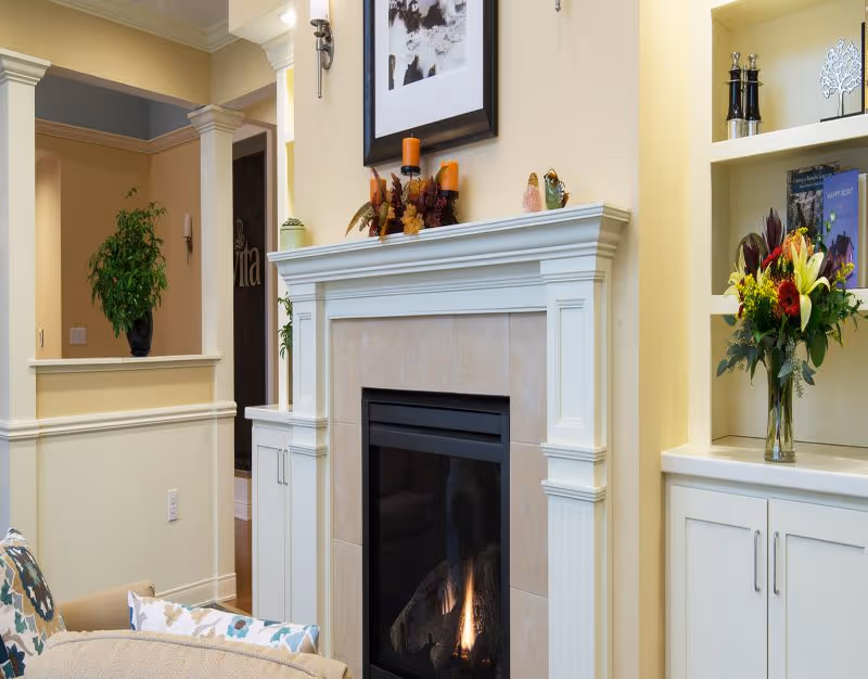 Cozy living room area with a lit fireplace surrounded by a white mantel. Above the mantel is a framed black and white picture and decorative candles. To the right, there is a built-in white cabinet with shelves holding a vase of fresh flowers and decorative items. A beige armchair with patterned cushions is partially visible in the foreground. The walls are painted a soft yellow, and there is a green potted plant on a ledge in the background.