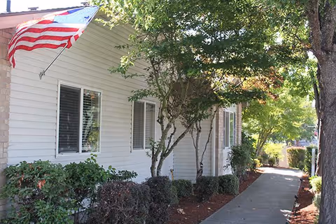 Side view of a white building with an American flag mounted near two windows. There are bushes and small trees along a concrete walkway that leads past the building, with sunlight filtering through the trees.