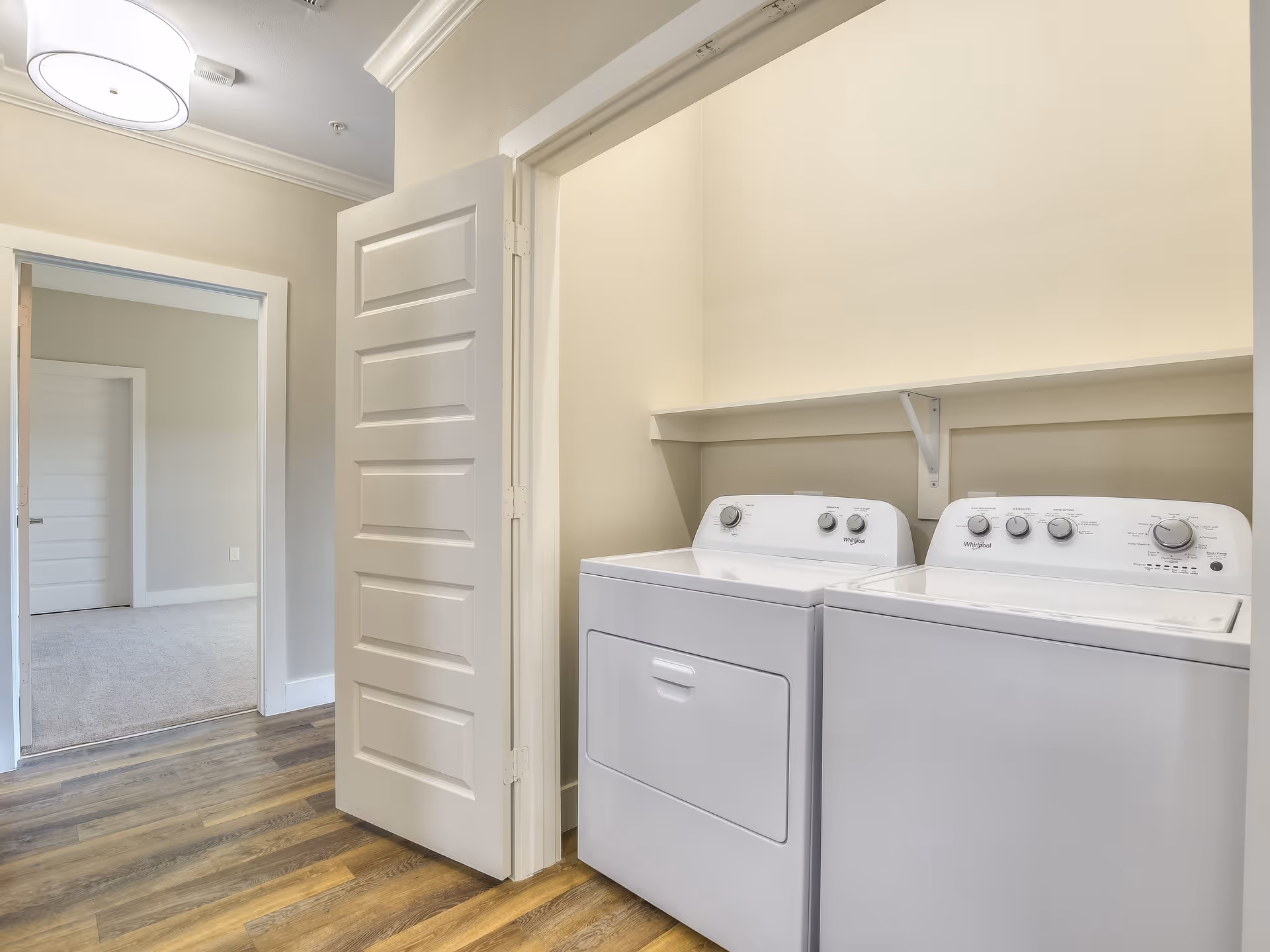 Interior view of a laundry area with a white washing machine and dryer side by side under a white shelf. The room has light beige walls and wood flooring. An open door leads to a carpeted room with light-colored walls.