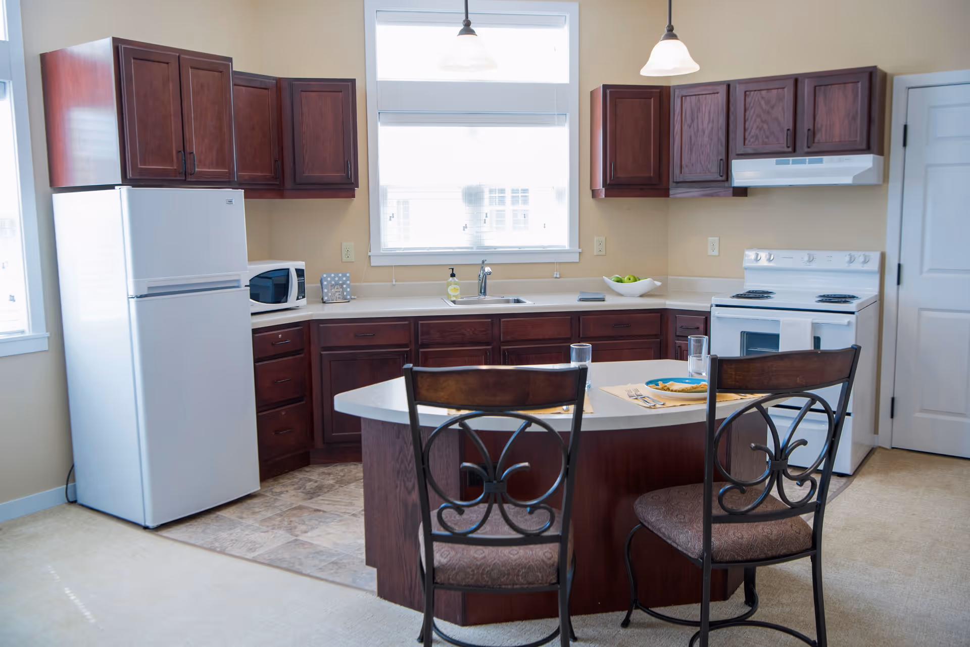 A bright kitchen with dark wooden cabinets, a white refrigerator, microwave, stove, and a central island with two chairs. The island has place settings with plates, glasses, and utensils. There is a window above the sink letting in natural light.