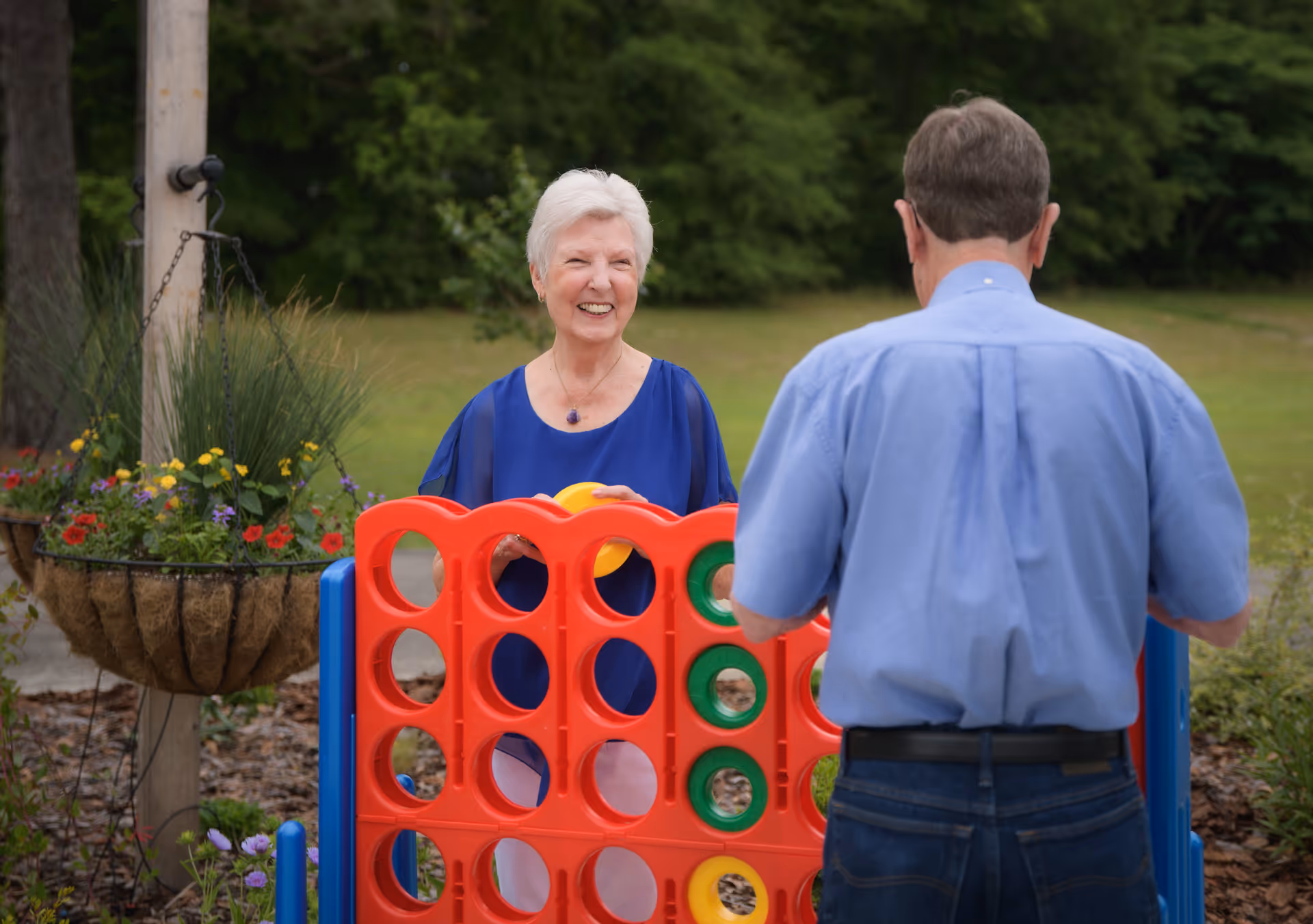 An elderly woman and man playing a large outdoor Connect Four game in a garden area with greenery and flowers around them. The woman is smiling and holding a yellow game piece, while the man is facing the game board.