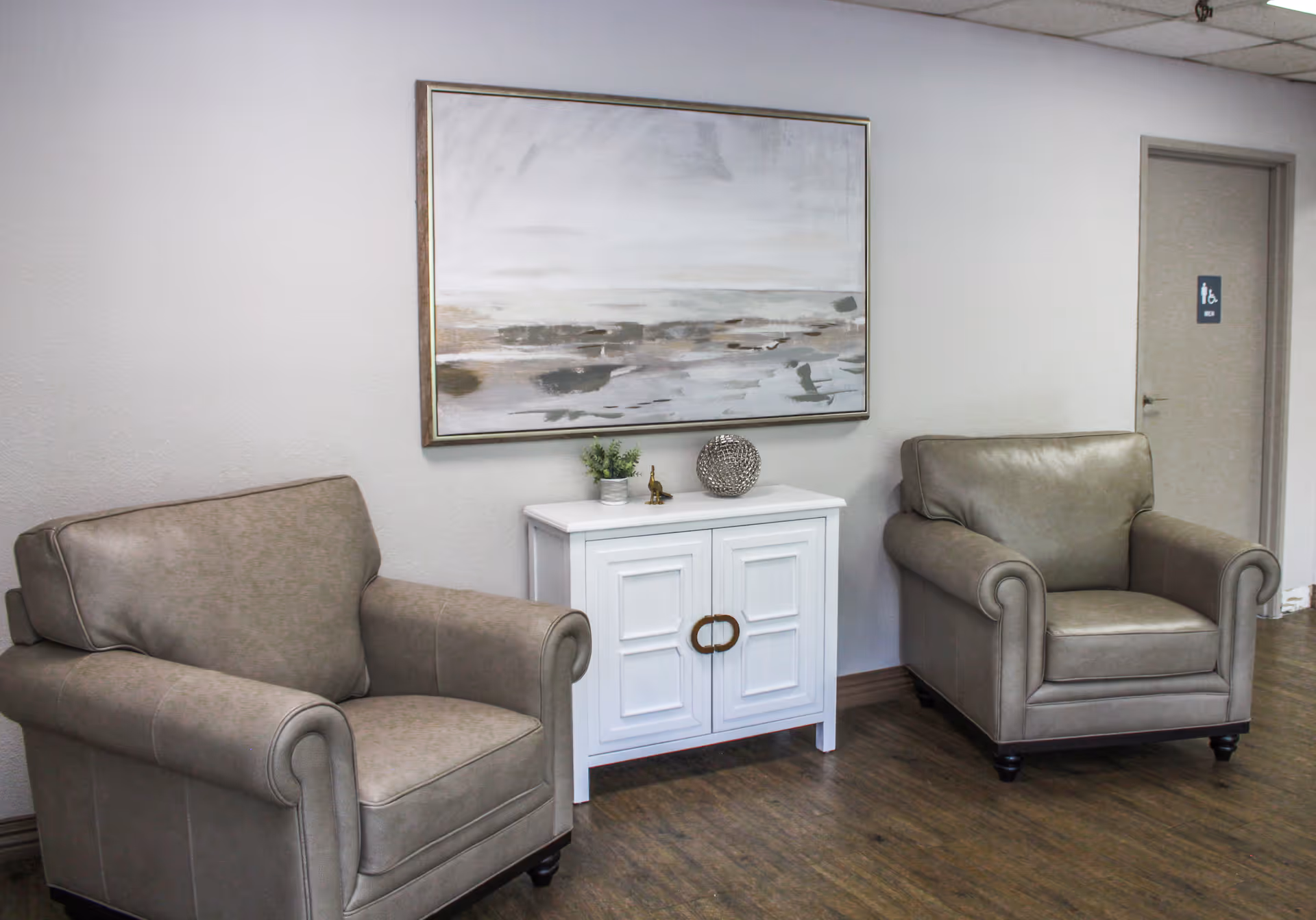 A seating area in a senior living facility with two beige armchairs placed on either side of a small white cabinet. On the cabinet are decorative items including a small plant and a spherical ornament. Above the cabinet hangs a large abstract painting with neutral tones. A door with a restroom sign is visible in the background.
