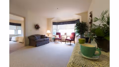 Bright living room with a sofa, two armchairs by a bay window, and a table with a green cup and potted plants in the foreground.