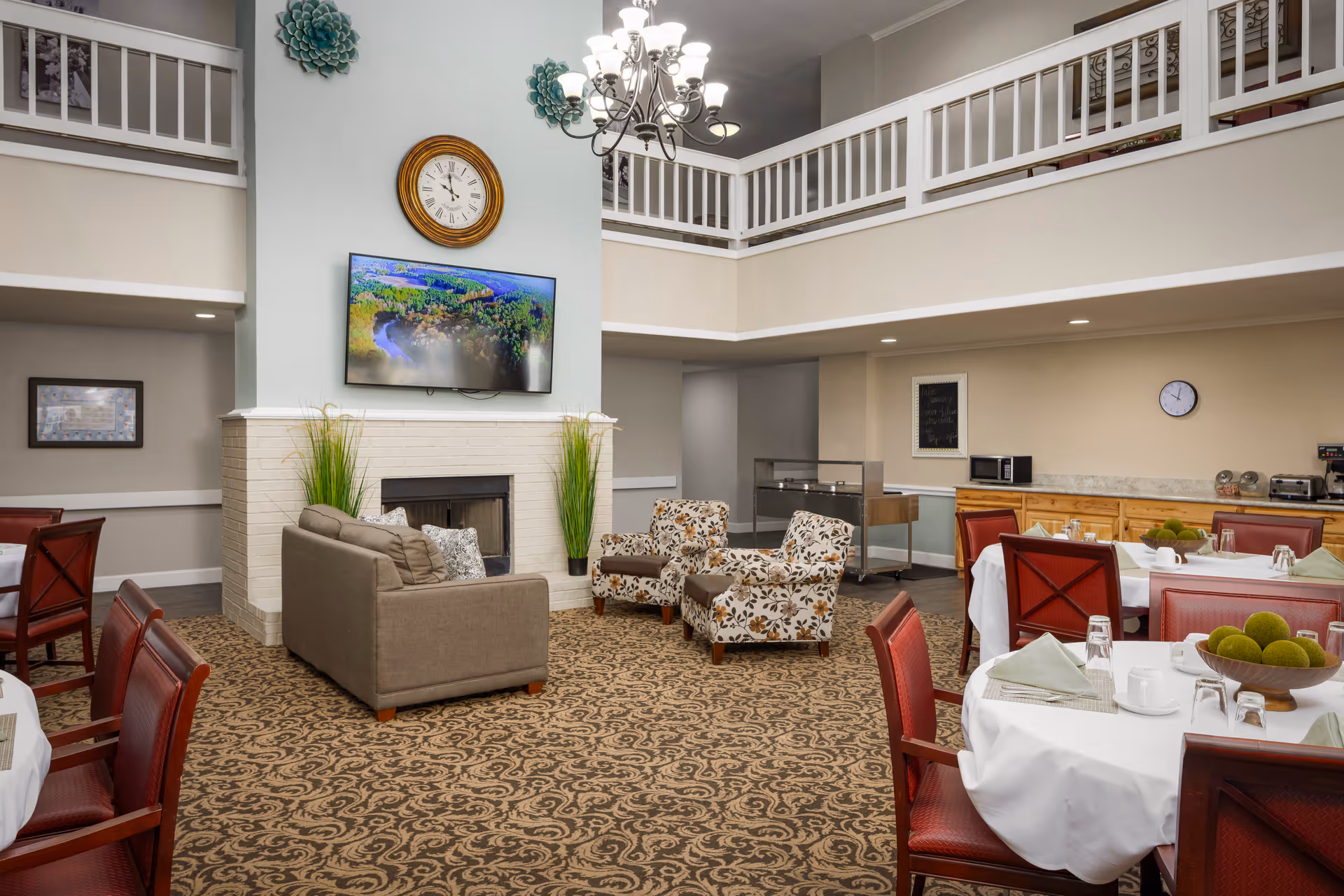 A spacious and well-lit dining and lounge area featuring round tables with white tablecloths and red chairs, set with cups, glasses, and napkins. In the center, there is a cozy seating area with a beige sofa and two floral-patterned armchairs arranged around a fireplace. Above the fireplace, a flat-screen TV and a round wall clock are mounted. The room has a high ceiling with a chandelier and a second-floor balcony railing visible.