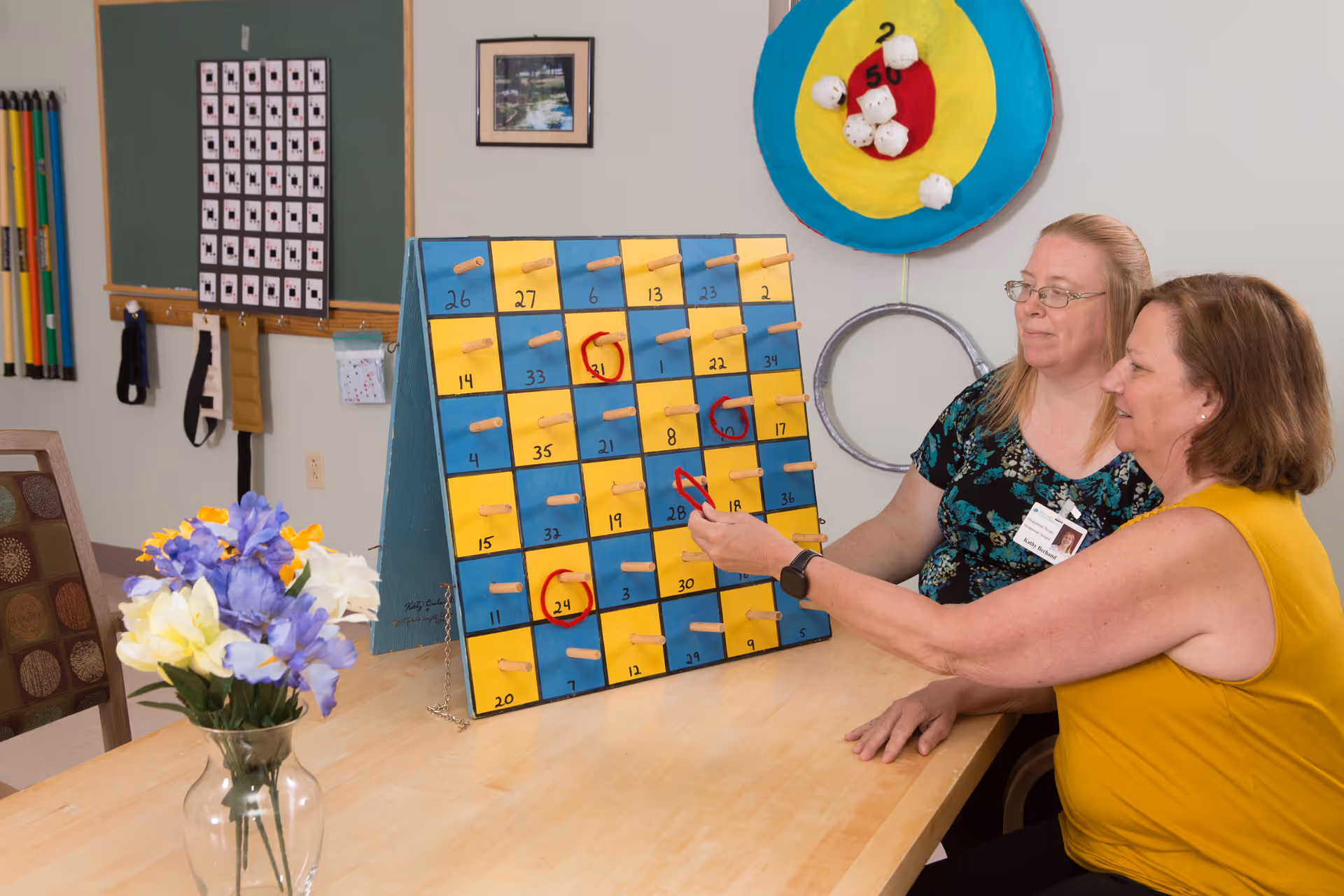 Two women sitting at a table playing a ring toss game with a large board featuring numbered squares and pegs. One woman is pointing at the board while the other watches. In the background, there is a colorful target with rings and a vase with purple and yellow flowers on the table.