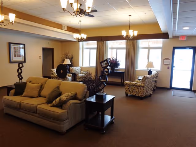 A spacious living room area in an assisted living facility with beige carpeting, a beige sofa with multiple cushions, two patterned armchairs, side tables with decorative sculptures, table lamps, and large windows with curtains allowing natural light to enter.
