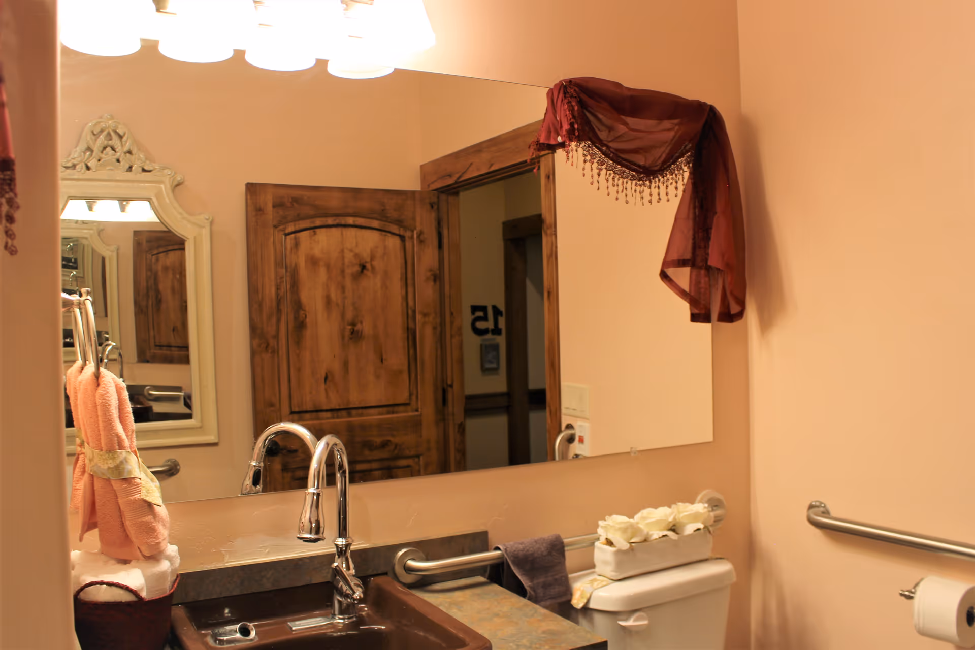 A bathroom with a brown sink and countertop, a large mirror above the sink, a wooden door reflected in the mirror, a toilet with a decorative white flower arrangement on the tank, grab bars on the walls, and peach-colored walls. There are pink towels hanging on a towel ring and a burgundy decorative cloth draped over the top corner of the mirror.