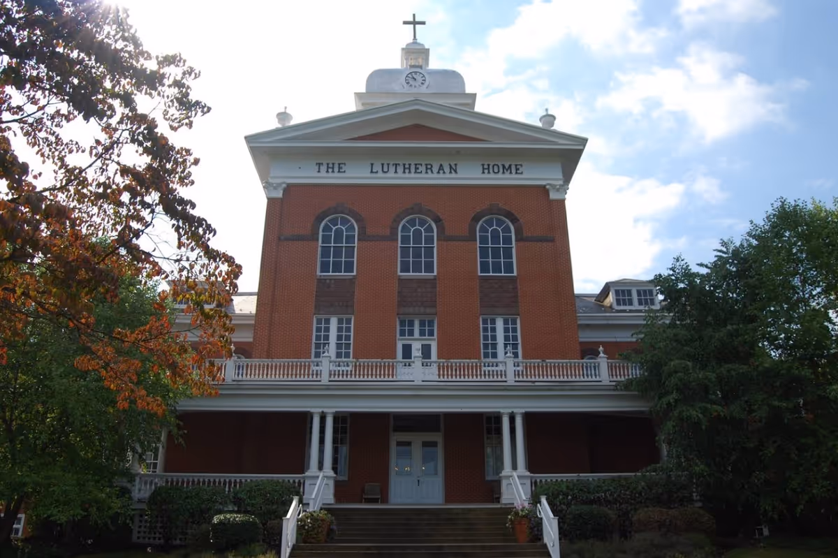 Front exterior view of a large brick building with white trim and a clock tower topped with a cross. The building has three tall arched windows and a sign that reads 'THE LUTHERAN HOME'. There are trees with green and some autumn-colored leaves on either side of the building.