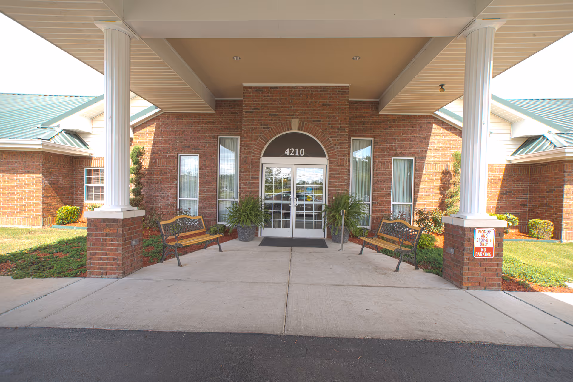 Covered entrance of a brick building with white columns, benches, potted plants, and double glass doors marked 4210.